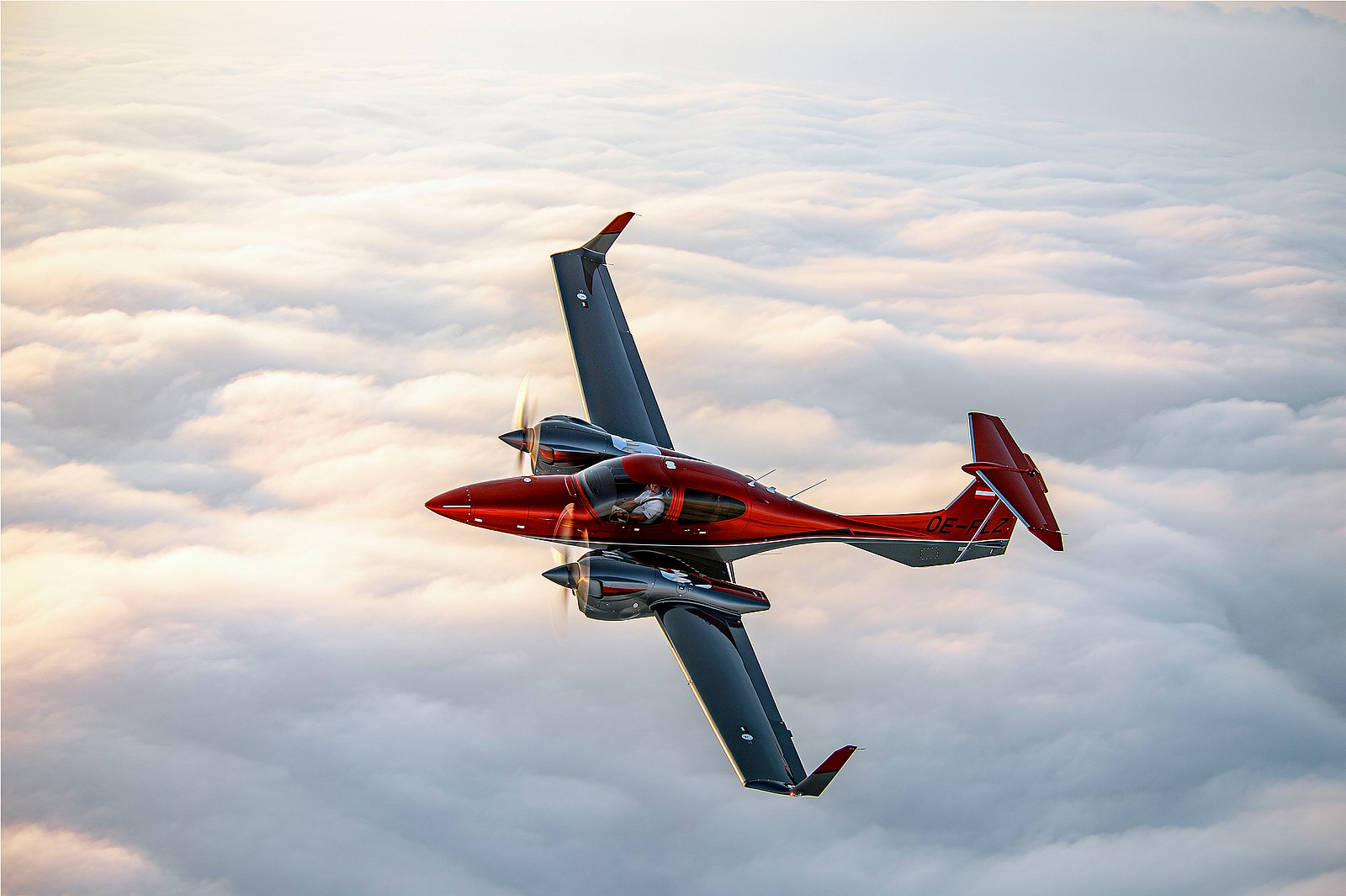 Switchblade aircraft in flight mode with retracted landing gear, red body reflecting golden sunlight over cloud layer.