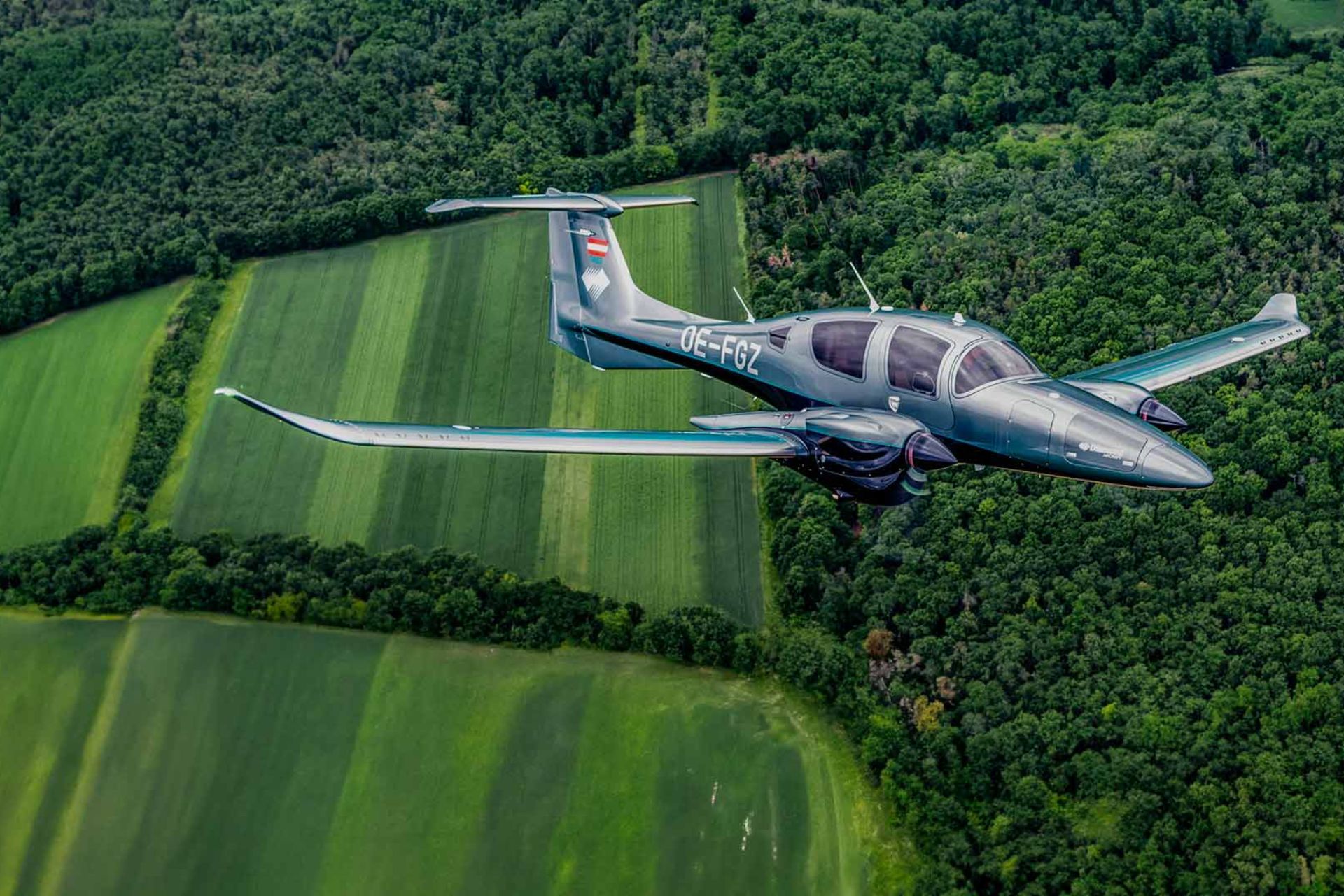 Diamond DA62 twin-engine aircraft OE-FGZ flying over lush green farmland and forest.
