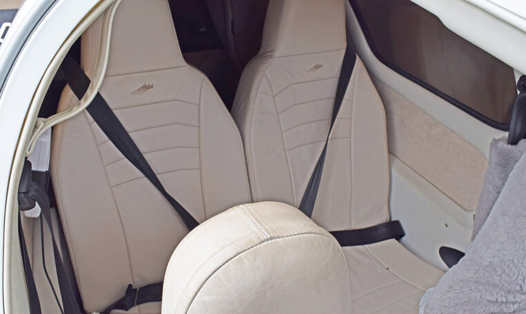 A view into the cabin of a small aircraft, showing two light beige leather passenger seats, each with a black diagonal seatbelt.