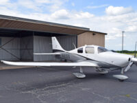 A white and grey single-engine propeller plane, registration number N603DG, parked on an asphalt tarmac in front of an open hangar, under a partly cloudy sky.