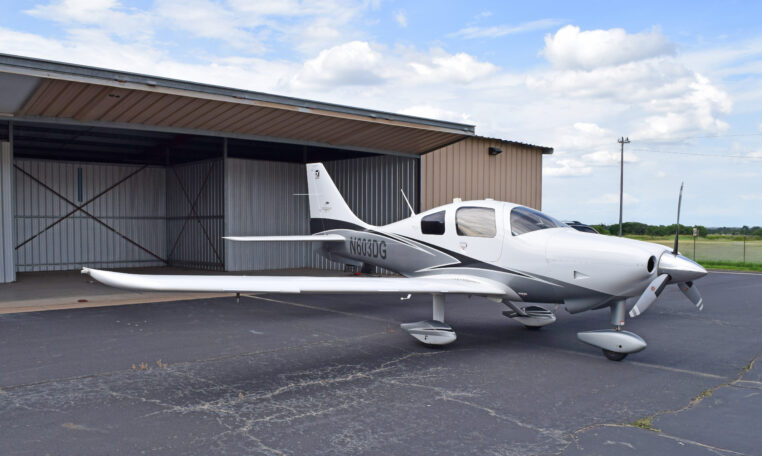 A white and grey single-engine propeller plane, registration number N603DG, parked on an asphalt tarmac in front of an open hangar, under a partly cloudy sky.