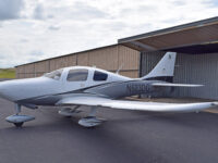 A white and grey single-engine propeller plane, registration number N603DG, parked on an asphalt tarmac in front of an open hangar, under a partly cloudy sky.
