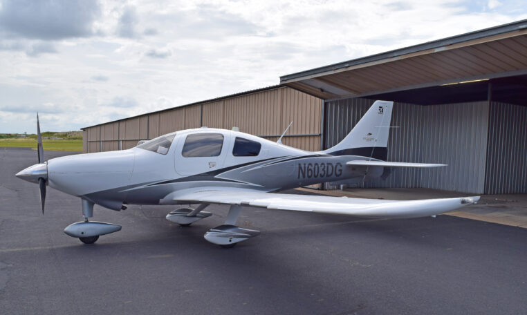 A white and grey single-engine propeller plane, registration number N603DG, parked on an asphalt tarmac in front of an open hangar, under a partly cloudy sky.
