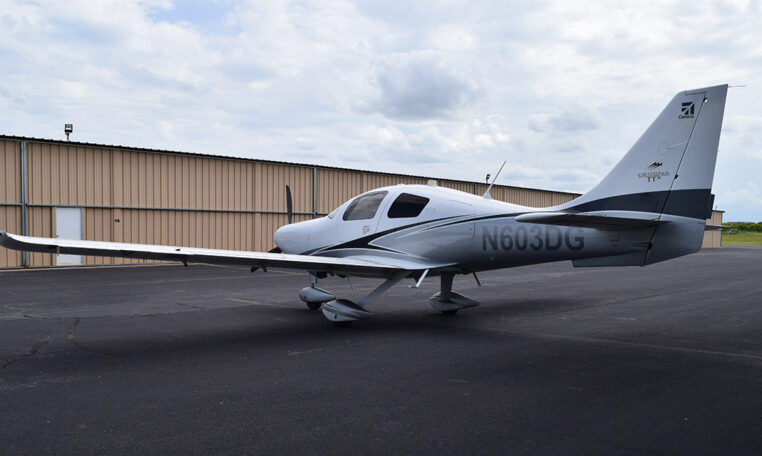 A white and grey single-engine propeller plane, registration number N603DG, parked on an asphalt tarmac in front of an open hangar, under a partly cloudy sky.