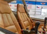 A side view of an aircraft cockpit, showing two tan leather pilot seats, dual control sticks, and an aircraft hangar visible in the background.