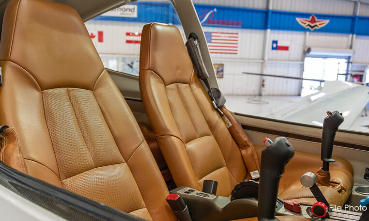 A side view of an aircraft cockpit, showing two tan leather pilot seats, dual control sticks, and an aircraft hangar visible in the background.