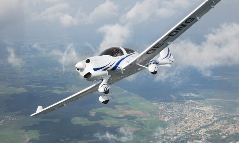 A white and blue single-engine propeller plane, registration OE-DNG, banking in flight high above a landscape of towns and fields.