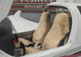 Light aircraft cockpit with sheepskin seats and dual controls under open canopy in hangar, Premier Aircraft sign.