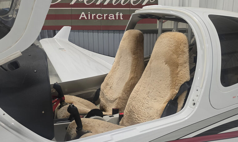 Light aircraft cockpit with sheepskin seats and dual controls under open canopy in hangar, Premier Aircraft sign.