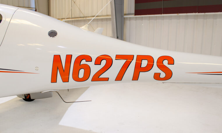 Close-up of white aircraft with orange tail number N627PS in a hangar, showing fuselage registration markings.