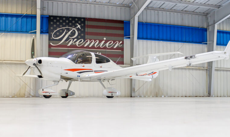 White light aircraft in hangar, side view of single-engine prop plane with orange accents under Premier sign.