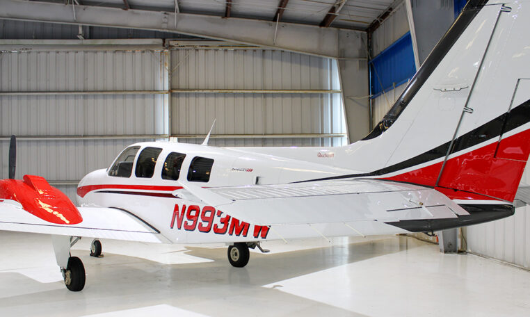 Beechcraft Bonanza single-engine airplane in a hangar, white with red accents, private aircraft side view.