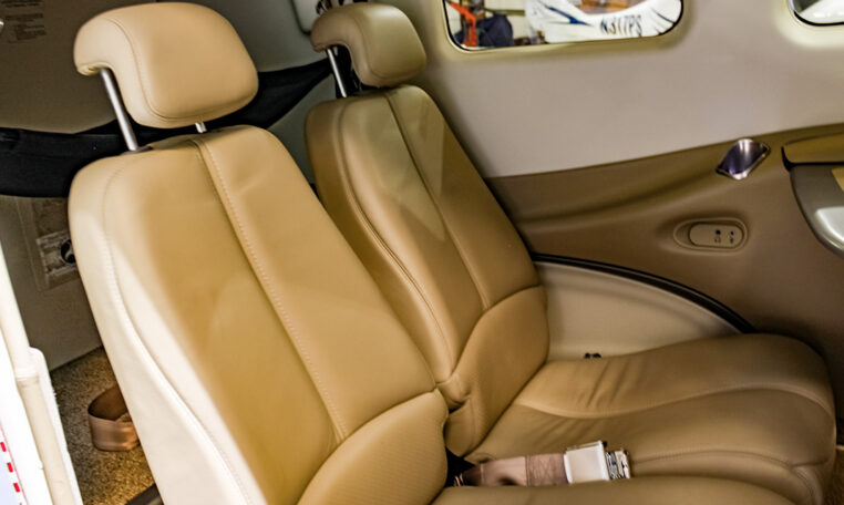 Tan leather passenger seats in a small private aircraft cabin, with headrests, seat belts and side window.