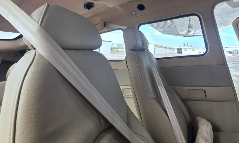 Small airplane interior with beige leather seats and fastened seatbelts, window view of airport hangars on tarmac.