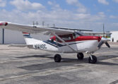 Red and white Cessna high-wing single-engine aircraft parked on airport tarmac by hangars