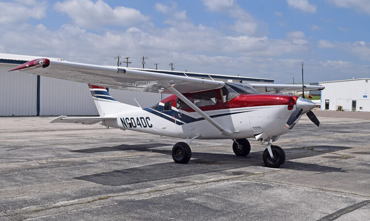 Red and white Cessna high-wing single-engine aircraft parked on airport tarmac by hangars