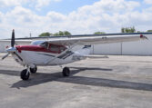 Cessna high-wing single-engine propeller plane, red and white, parked on airport tarmac in front of hangars under blue sky
