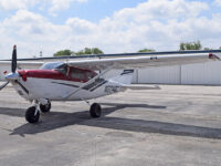 Cessna high-wing single-engine propeller plane, red and white, parked on airport tarmac in front of hangars under blue sky