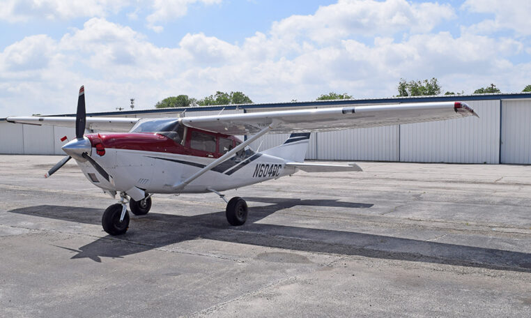 Cessna high-wing single-engine propeller plane, red and white, parked on airport tarmac in front of hangars under blue sky