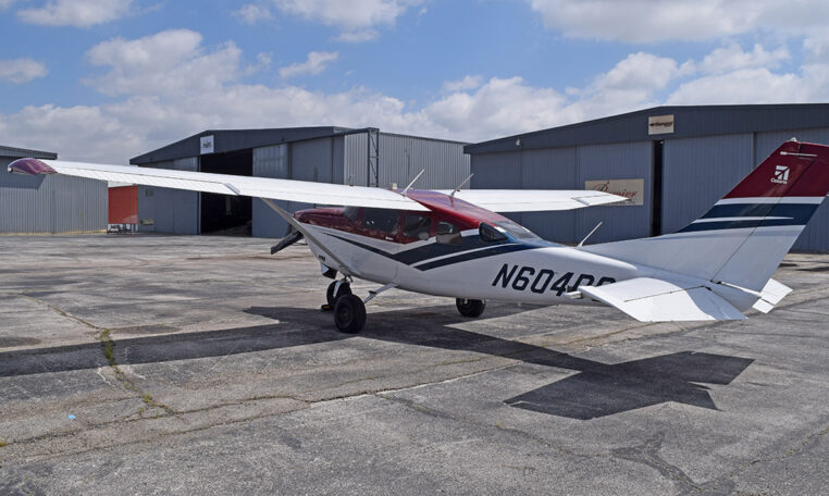 High-wing single-engine taildragger general aviation airplane parked by hangars on tarmac under blue sky