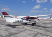 High-wing Cessna single-engine propeller plane parked on airport tarmac by hangars under a blue sky