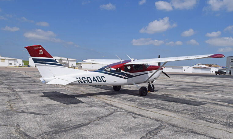 High-wing Cessna single-engine propeller plane parked on airport tarmac by hangars under a blue sky