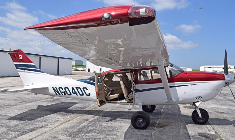 Red and white Cessna high-wing single-engine airplane on airport ramp, cabin door open for boarding.