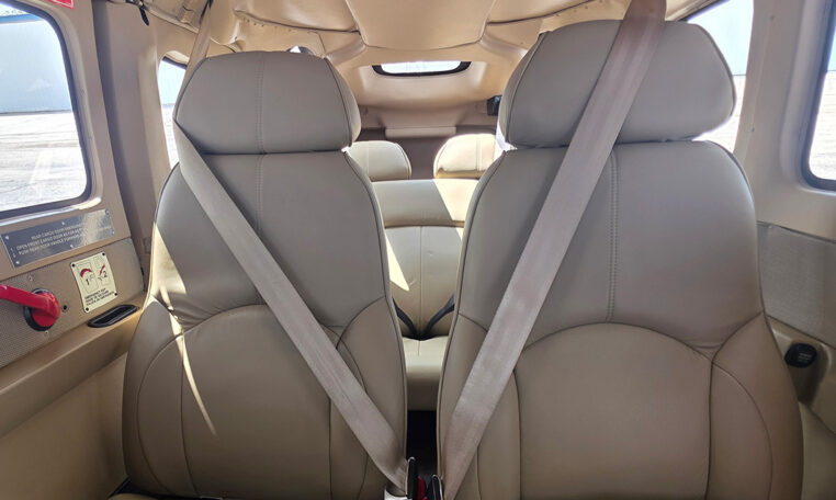 Private plane cabin interior with tan leather seats and seatbelts; rear bench visible at airport.