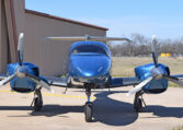Blue twin-engine propeller aircraft parked on airport tarmac by hangar, front view on a sunny day.