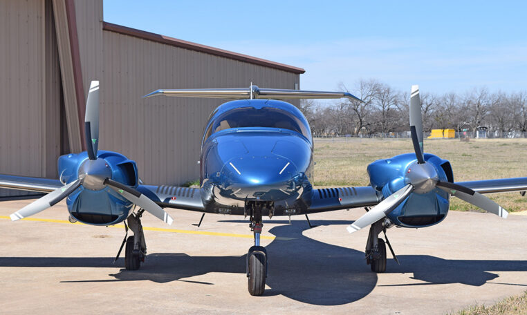 Blue twin-engine propeller aircraft parked on airport tarmac by hangar, front view on a sunny day.