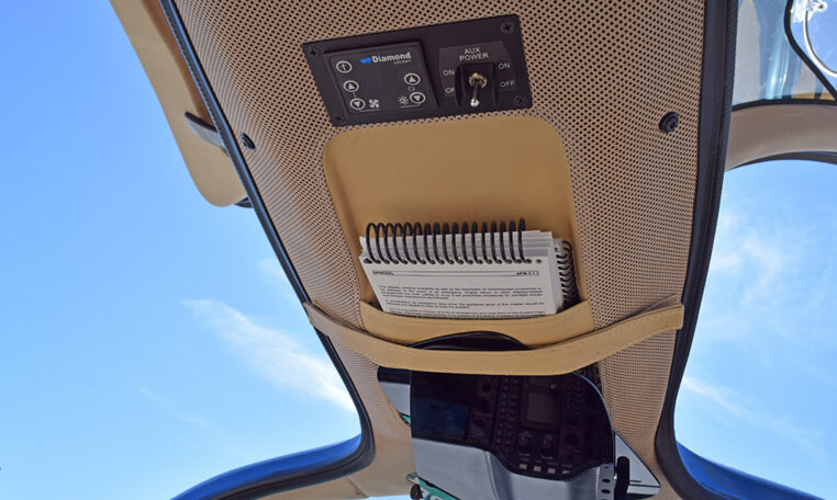 Diamond Aircraft cockpit overhead panel with AUX power controls and pilot checklist pocket, blue sky view.