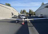 Single-engine light aircraft parked on airport apron between steel hangars, front view under blue sky