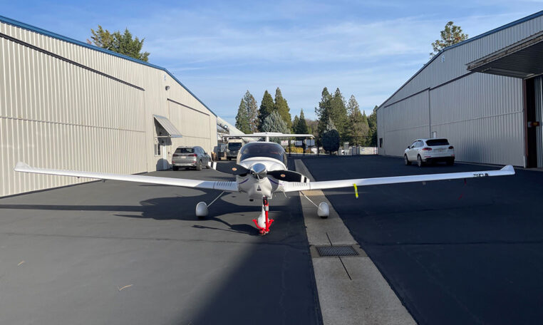 Single-engine light aircraft parked on airport apron between steel hangars, front view under blue sky