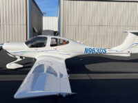 White single-engine light airplane N963DS parked on tarmac beside metal hangars under blue sky.