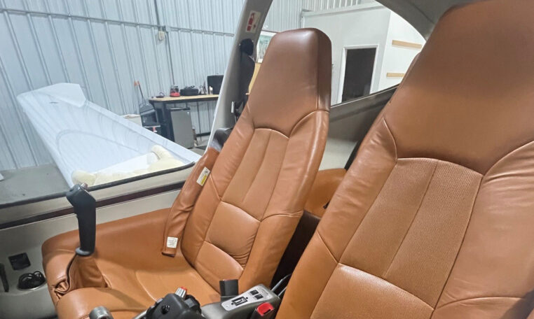 Airplane cockpit interior with two tan leather seats and joystick controls inside a hangar