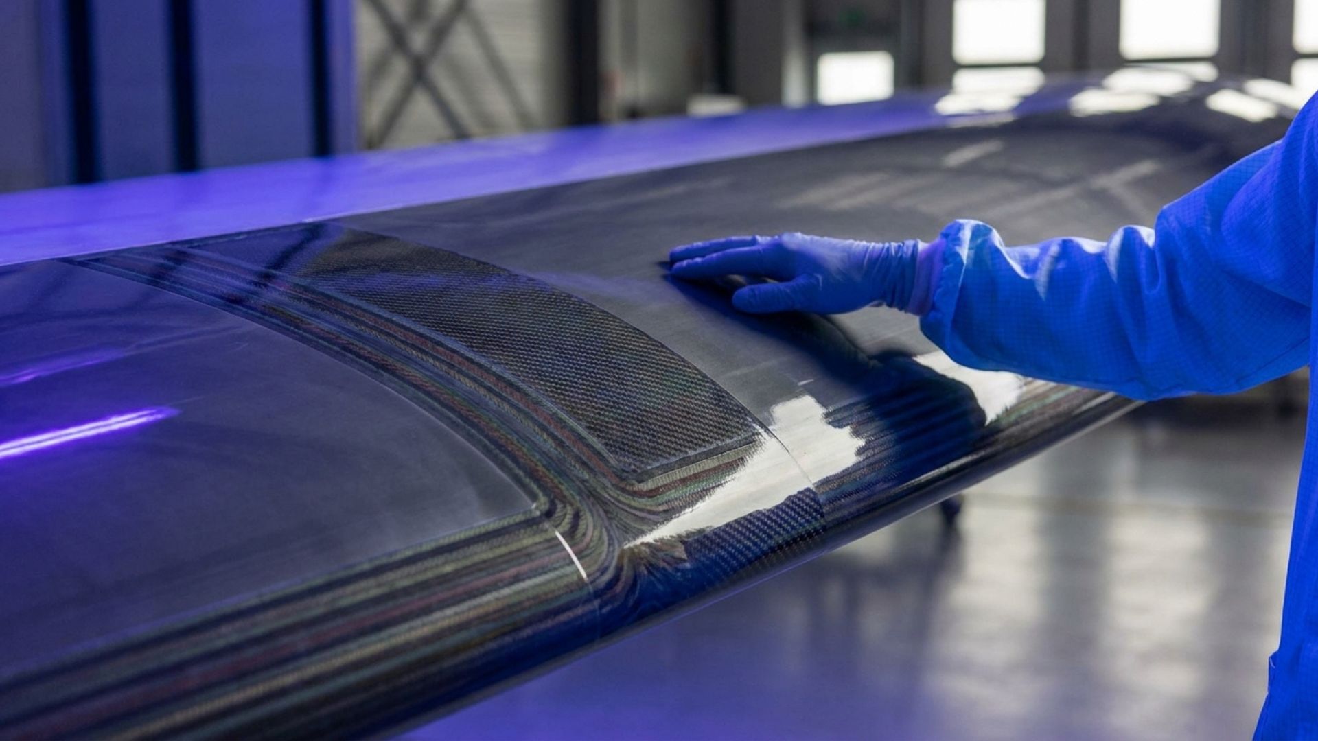 Gloved technician inspecting carbon fiber aerospace wing surface in cleanroom, composite manufacturing quality control.