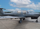 Diamond DA50 RG single-engine prop airplane in grey livery parked on airport tarmac under blue sky with clouds