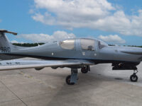 Diamond DA50 RG single-engine prop airplane in grey livery parked on airport tarmac under blue sky with clouds