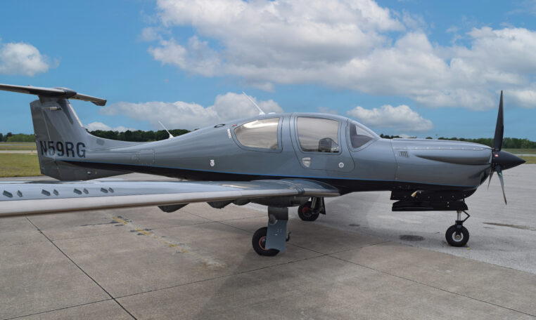Diamond DA50 RG single-engine prop airplane in grey livery parked on airport tarmac under blue sky with clouds