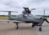 Sleek gray single-engine propeller airplane on airport tarmac with gullwing cockpit doors open, ready for flight