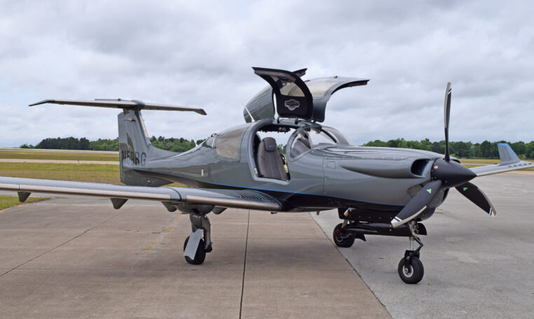 Sleek gray single-engine propeller airplane on airport tarmac with gullwing cockpit doors open, ready for flight