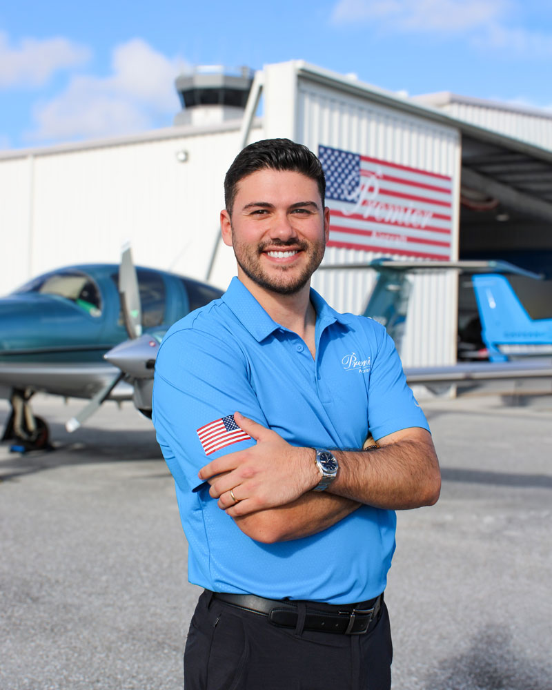 Michael Smelcer. Smiling pilot in blue polo at aircraft hangar with American flag, small airplane in background at airport.