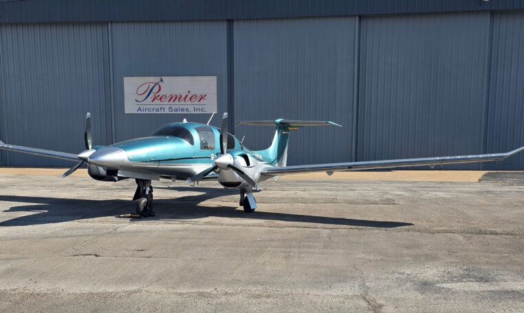 Teal and silver twin-engine private propeller airplane on tarmac at Premier Aircraft Sales hangar, front view