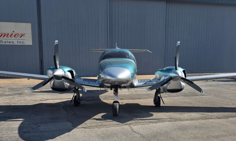 Front view of a teal-silver twin-engine propeller aircraft with three-blade props on tarmac outside a hangar
