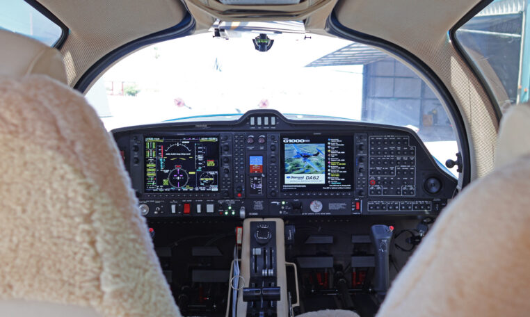 Diamond DA62 glass cockpit with Garmin G1000 NXi avionics and dual yokes, view from cabin seats in hangar.