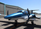 Sleek blue twin-engine propeller private airplane parked on sunny airport tarmac beside hangars