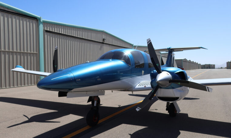 Sleek blue twin-engine propeller private airplane parked on sunny airport tarmac beside hangars