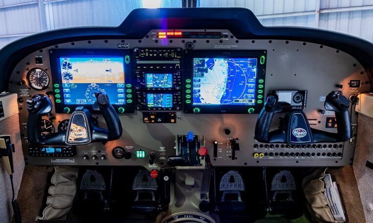 Light aircraft cockpit with Garmin G1000 glass avionics, dual yokes, PFD and MFD navigation displays in a hangar.