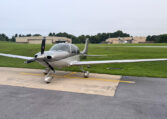 Silver single-engine propeller airplane parked on the tarmac at a small airport with hangars in the background.