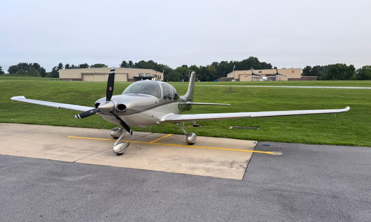 Silver single-engine propeller airplane parked on the tarmac at a small airport with hangars in the background.